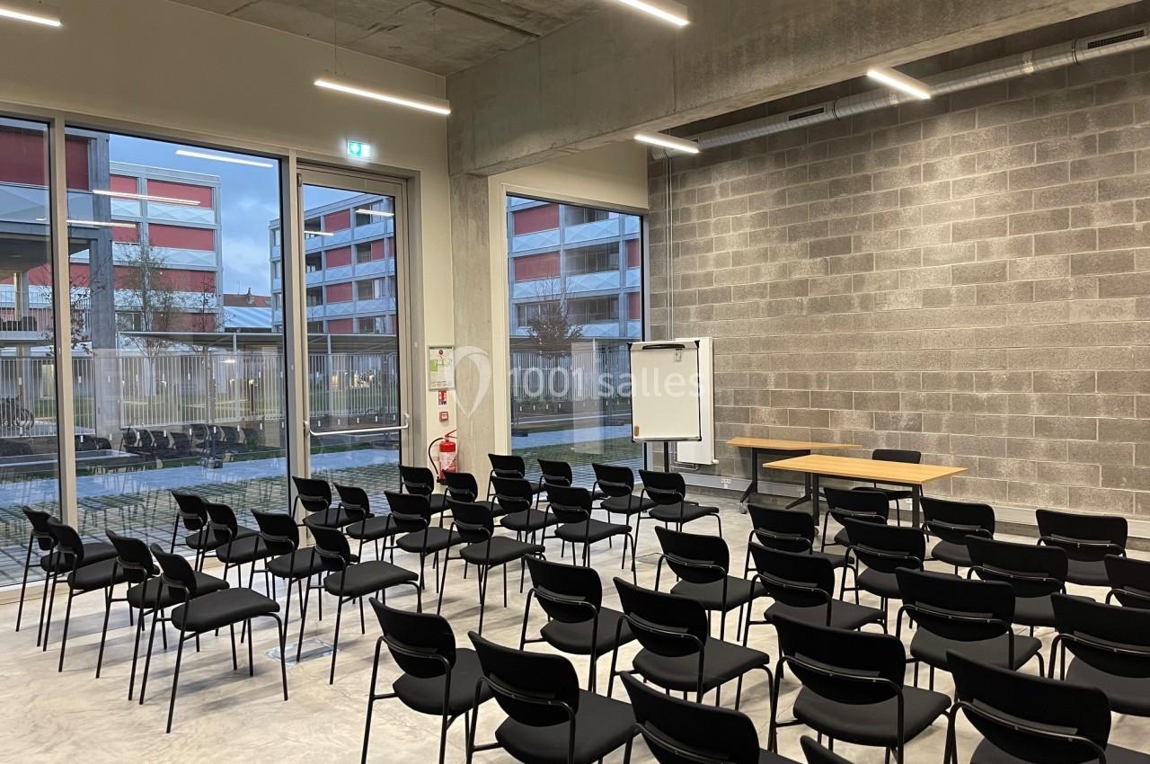 Salle de conférence vide avec des rangées de chaises noires, un tableau blanc et une table, éclairée par des fenêtres.