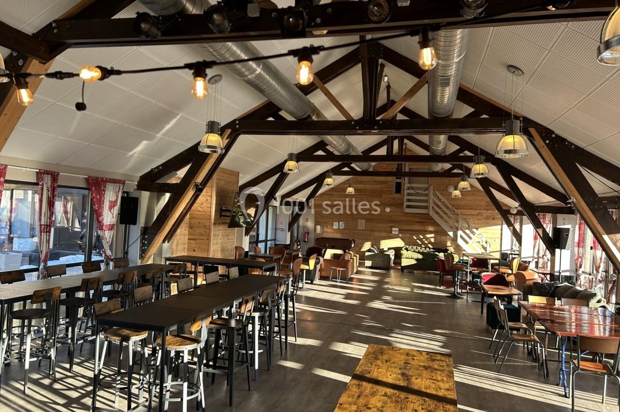 Salle spacieuse avec charpente en bois apparente, tables et chaises disposées pour accueillir des convives.