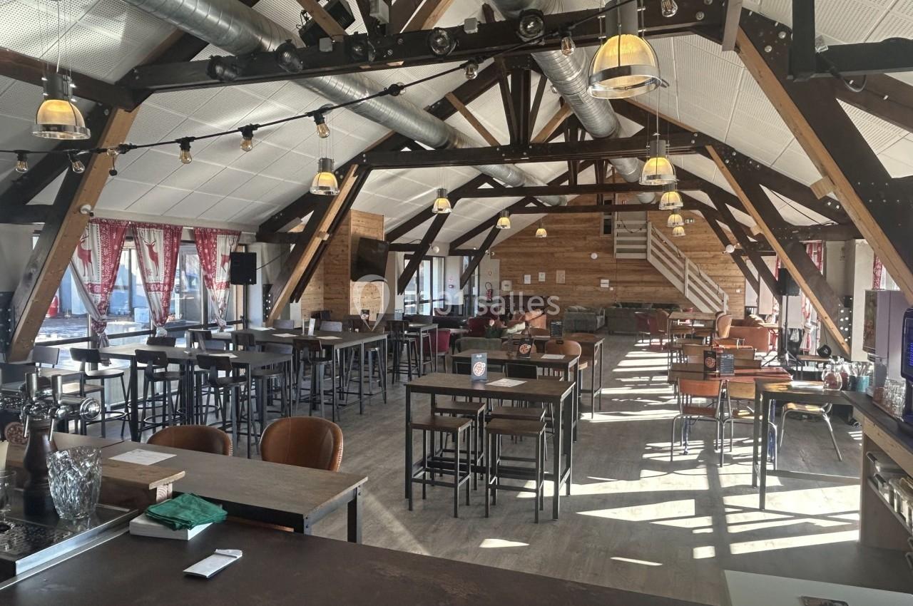 Grande salle lumineuse avec charpente en bois apparente, tables hautes, chaises et baies vitrées avec rideaux rouges.