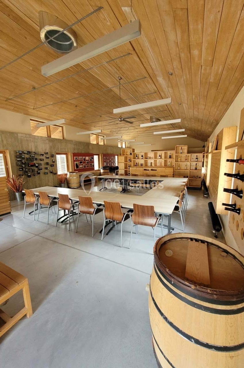 Salle de réunion lumineuse avec tables en U, chaises en bois, tonneau décoratif et étagères murales garnies de bouteilles.