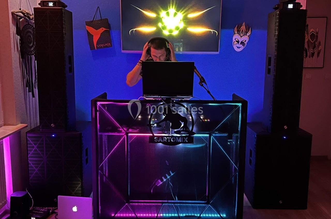 Un DJ mixe de la musique derrière une console lumineuse dans une pièce décorée de masques et d'enceintes.