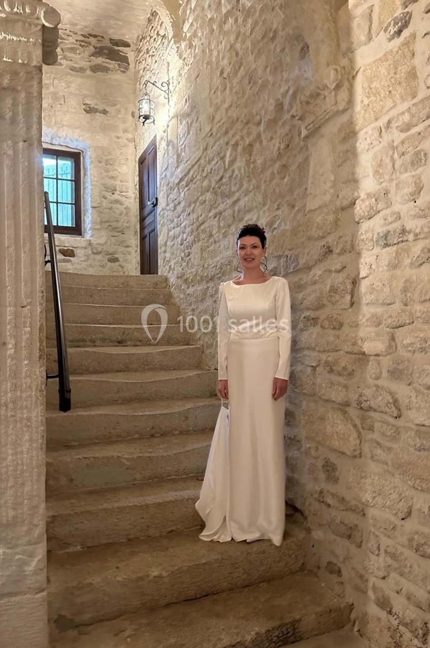 Femme en robe blanche debout près d'un escalier en pierre dans un intérieur ancien aux murs en pierre apparente.