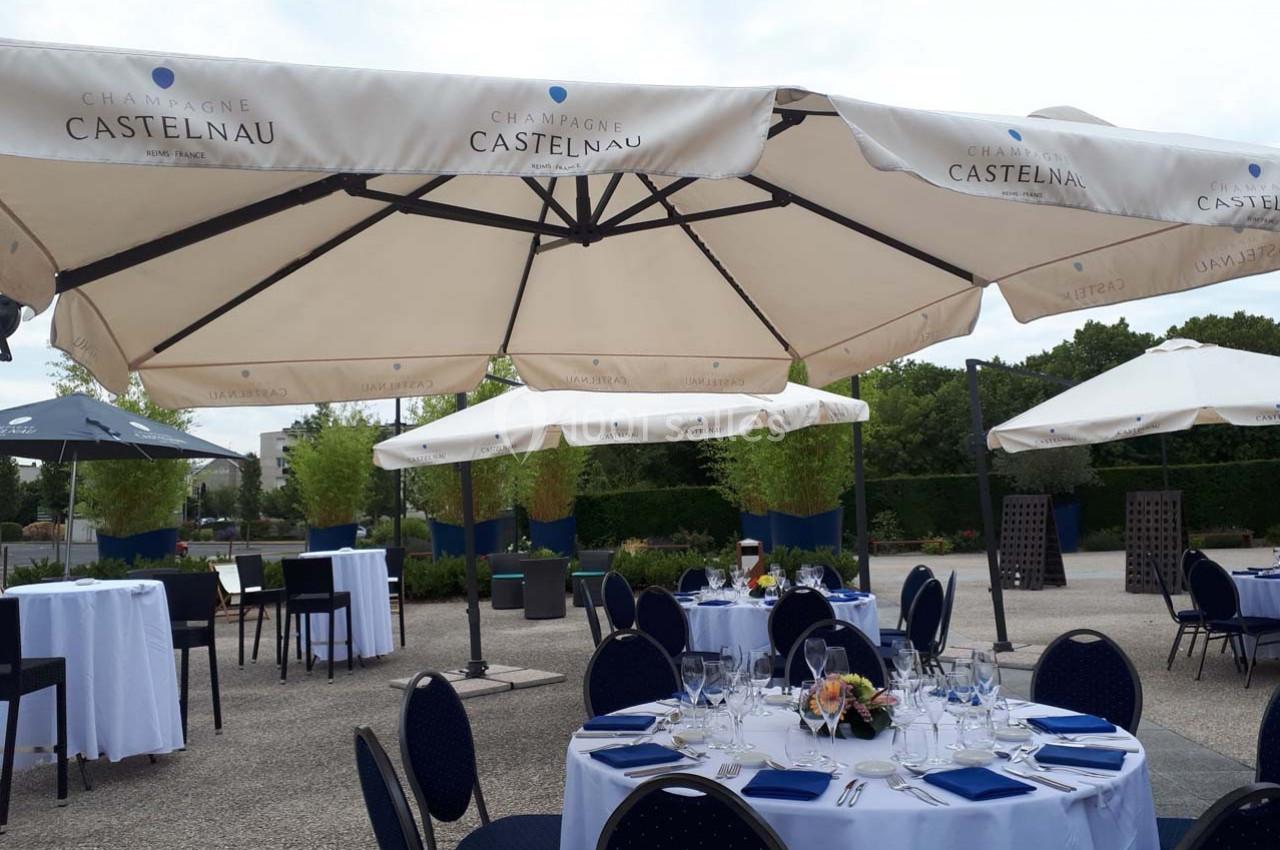 Tables dressées avec nappes blanches et chaises bleues sous de grands parasols dans un espace extérieur arboré.