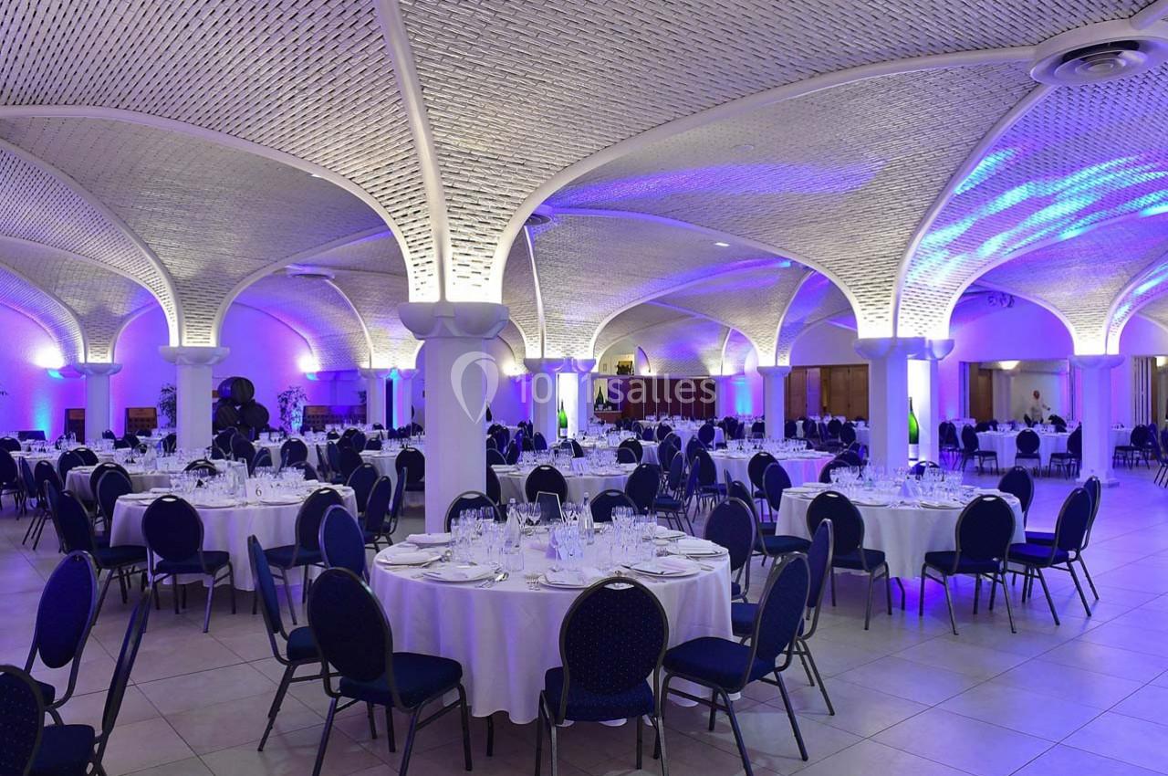 Salle de réception élégante avec tables rondes dressées, chaises bleues et éclairage violet sous des voûtes en pierre.