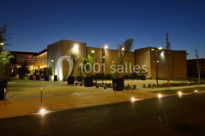 Bâtiment moderne éclairé de nuit, entouré de plantes et d'un espace extérieur aménagé avec des lampadaires.