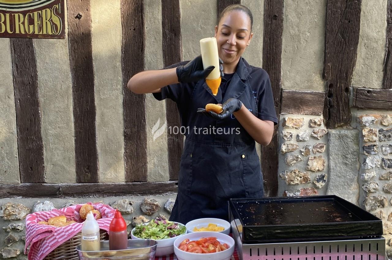 Une cuisinière prépare un burger en ajoutant une sauce, devant une table garnie d'ingrédients et une plaque de cuisson.
