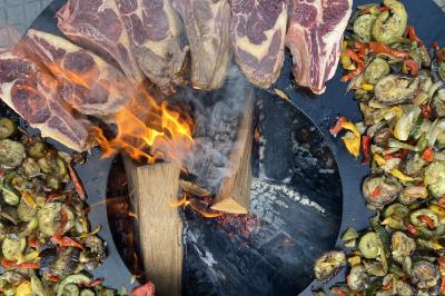 Deux hommes cuisinent des aliments sur un grand barbecue en plein air, éclairé par les flammes, à la tombée de la nuit.