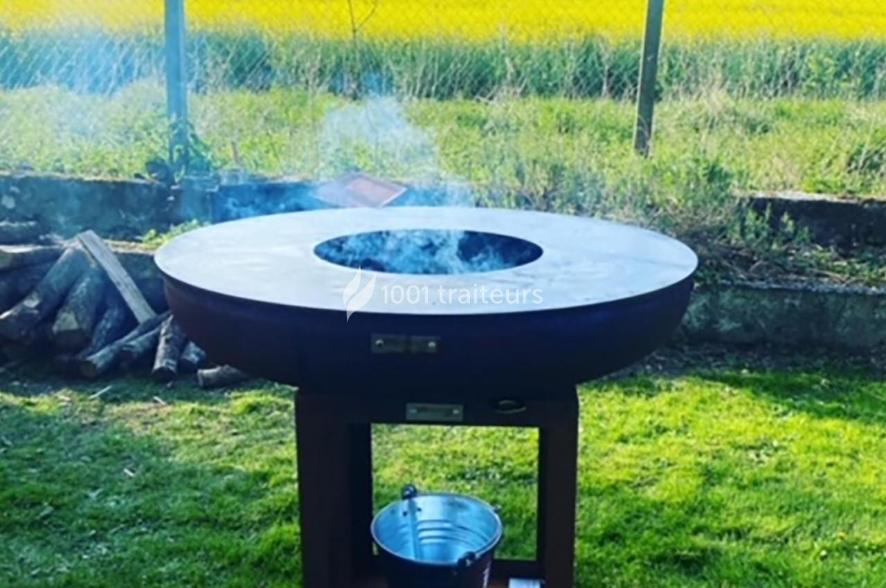 Barbecue rond en métal fumant, installé dans un jardin verdoyant avec des bûches empilées à proximité.