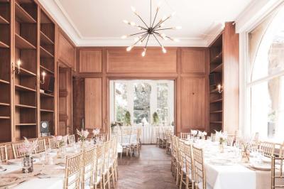 Salle lumineuse décorée pour un événement, avec des tables dressées, des chaises dorées et des fleurs en centre de table.