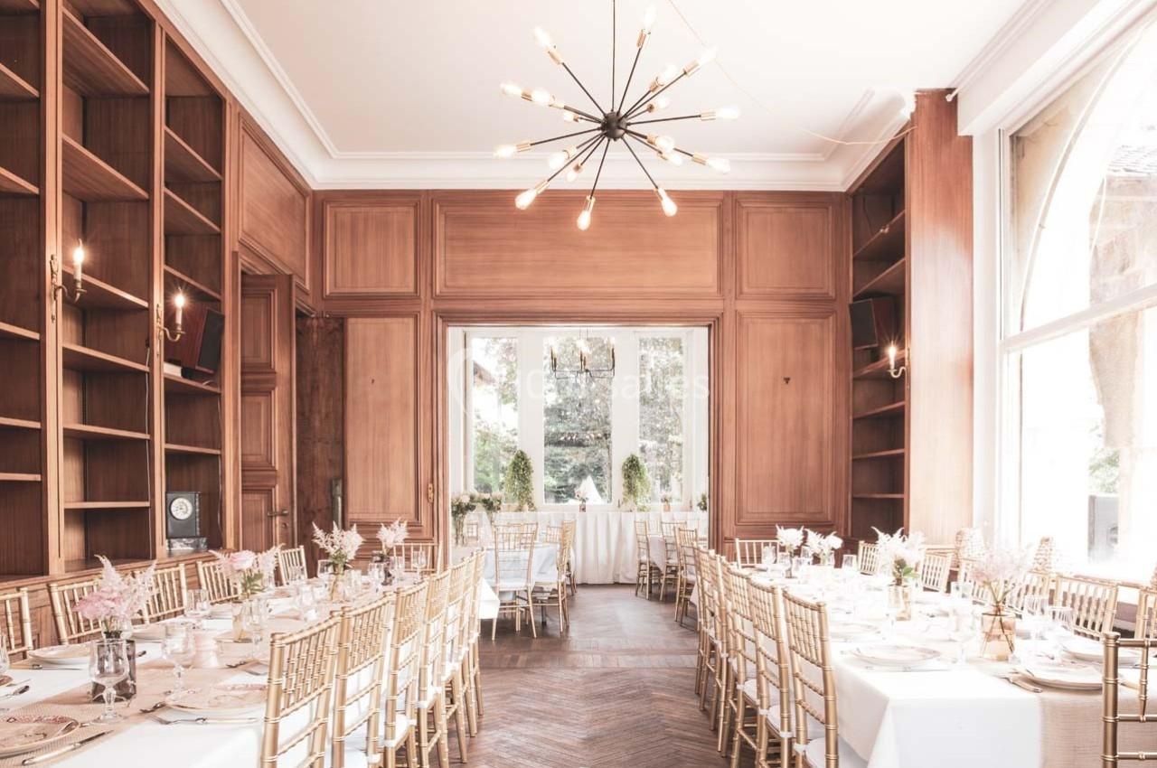Salle de réception lumineuse avec tables décorées, chaises dorées et grandes fenêtres donnant sur un jardin.
