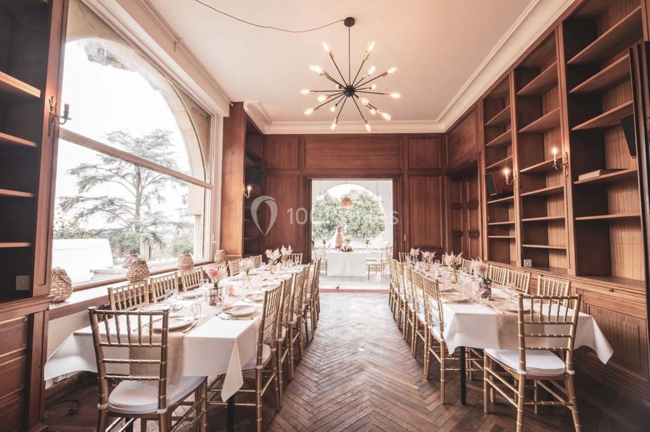 Salle à manger élégante avec de longues tables dressées, chaises dorées et grandes fenêtres donnant sur un jardin.