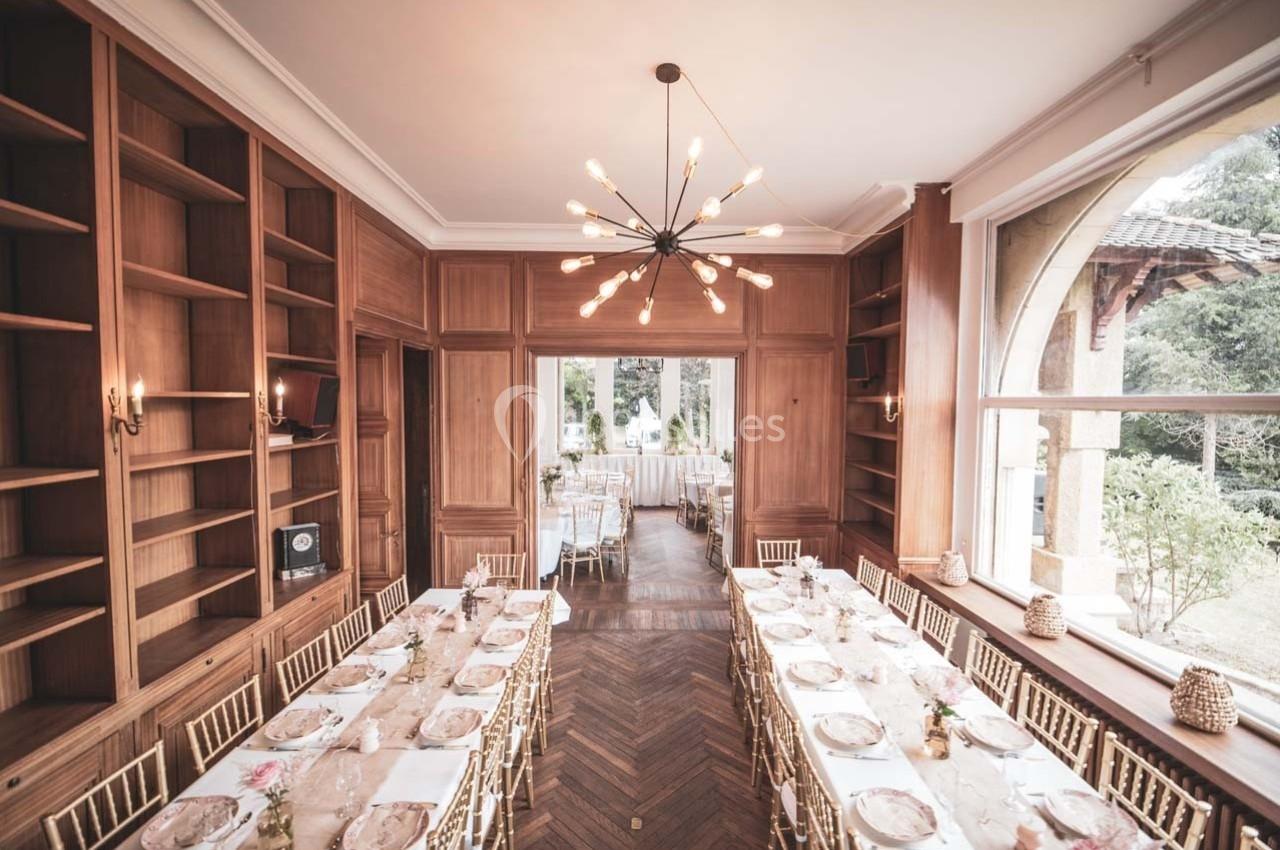 Salle élégante avec tables dressées, chaises dorées, parquet en bois, étagères et grande baie vitrée donnant sur un jardin.
