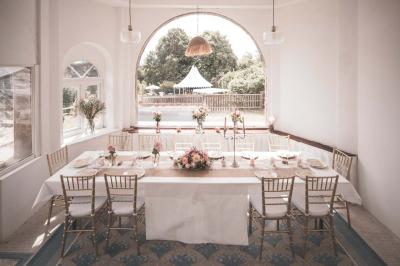 Salle lumineuse décorée pour un événement, avec des tables dressées, des chaises dorées et des fleurs en centre de table.
