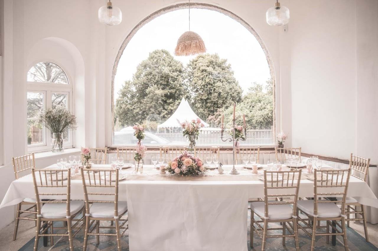 Table élégamment dressée dans une salle lumineuse avec de grandes fenêtres et une vue sur des arbres et une tente blanche.