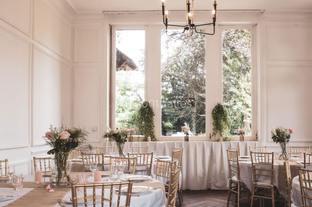 Salle lumineuse décorée pour un événement, avec des tables dressées, des chaises dorées et des fleurs sur les tables.