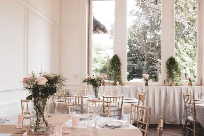 Salle lumineuse décorée pour un événement, avec des tables dressées, des chaises dorées et des fleurs en centre de table.