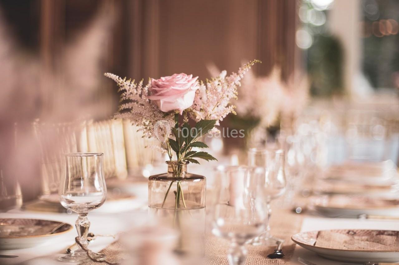 Table décorée avec des assiettes, verres et un vase contenant une rose rose et des fleurs délicates au centre.