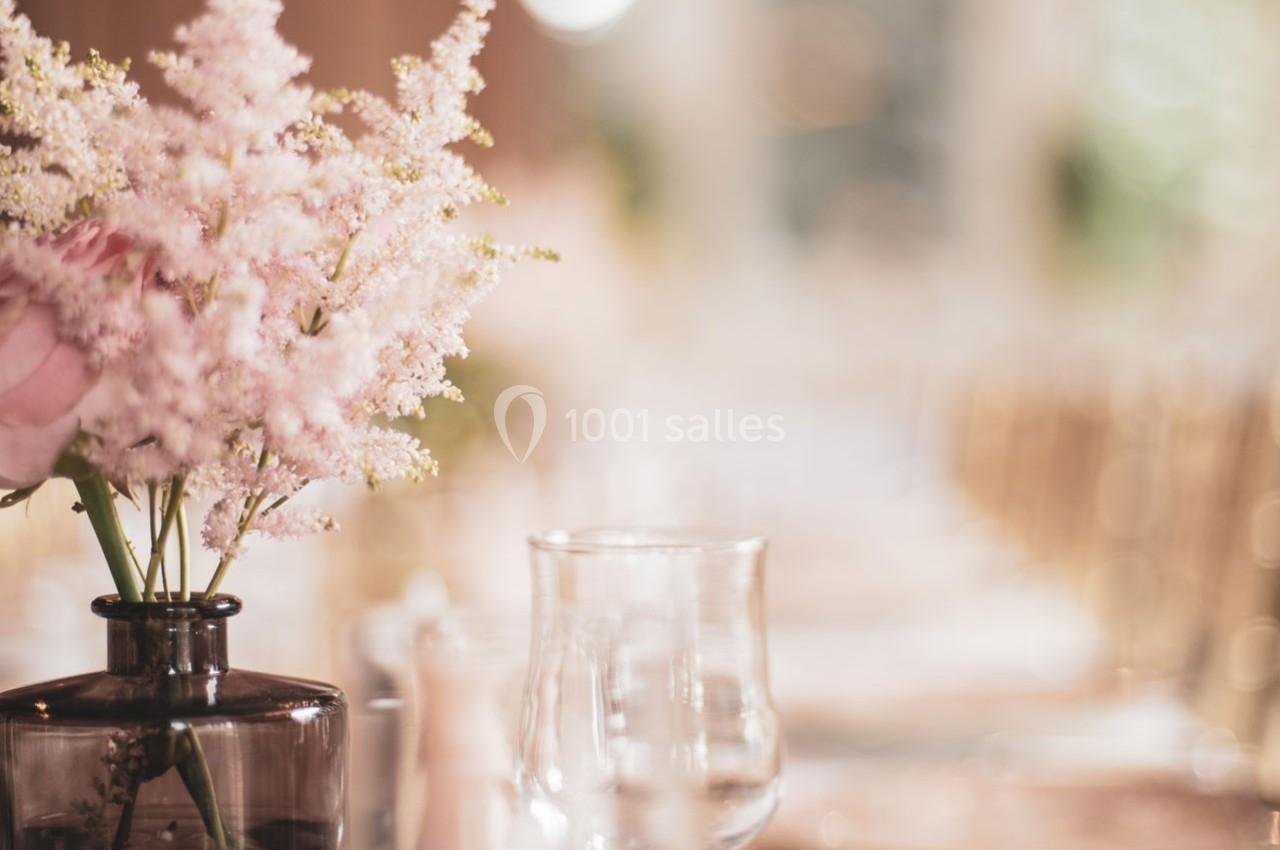 Vase en verre brun avec des fleurs roses délicates sur une table dressée dans un décor lumineux et flou.