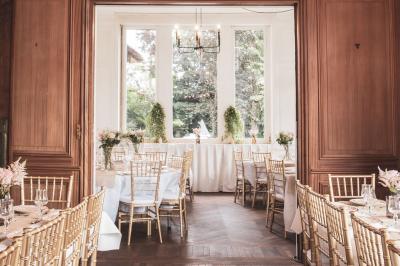 Salle lumineuse décorée pour un événement, avec des tables dressées, des chaises dorées et des fleurs en centre de table.