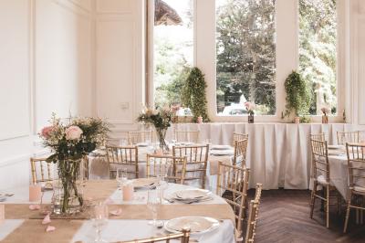 Salle lumineuse décorée pour un événement, avec des tables dressées, des chaises dorées et des fleurs en centre de table.