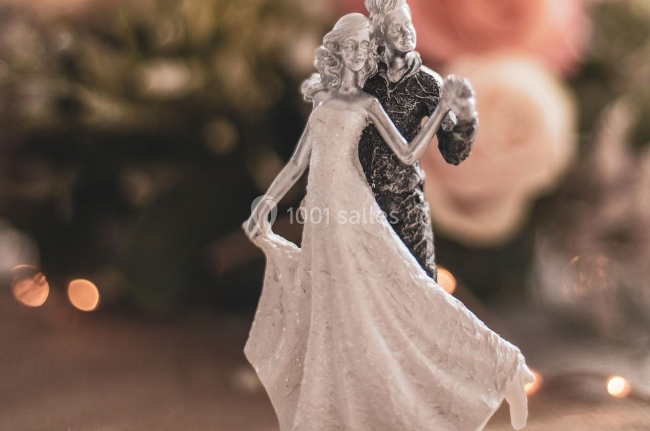 Figurine de mariés dansant, avec une robe de mariée fluide et un costume, sur un fond flou de fleurs et lumières.