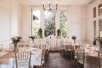 Salle lumineuse décorée pour un événement, avec des tables dressées, des chaises dorées et des fleurs en centre de table.