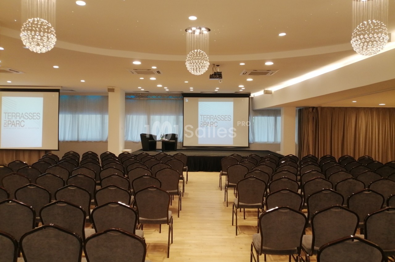 Salle de conférence avec rangées de chaises vides, deux écrans de projection et éclairage suspendu.