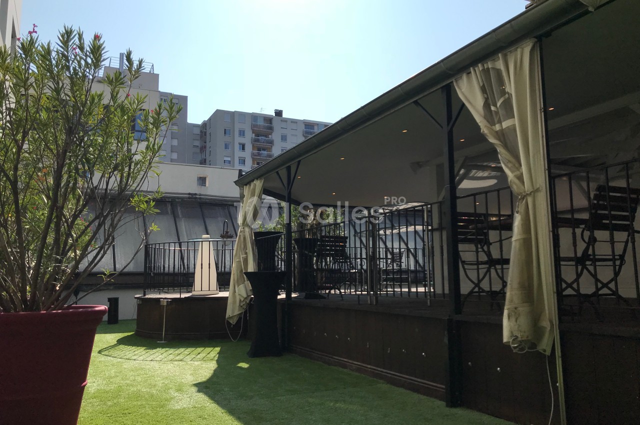 Terrasse extérieure avec pelouse artificielle, pergola couverte et immeubles en arrière-plan par temps ensoleillé.