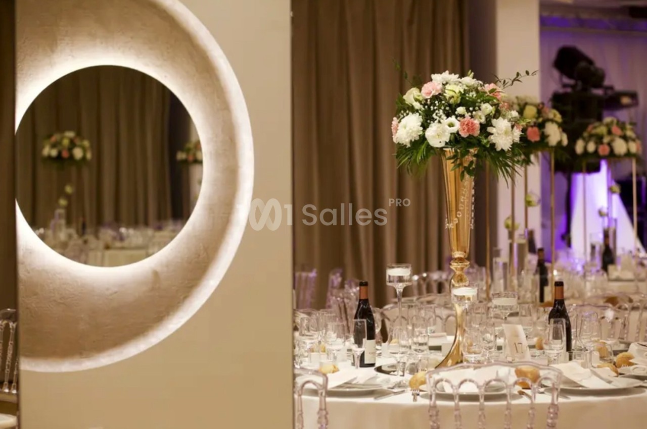 Salle de réception élégante avec tables dressées, décor floral sur pied doré et éclairage tamisé.