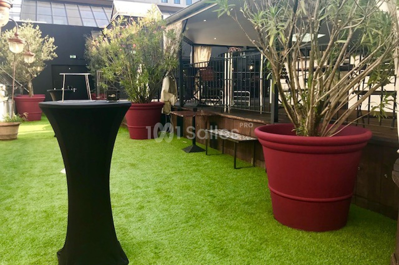 Terrasse avec gazon artificiel, grandes plantes en pots rouges et mobilier de réception près d'une estrade.