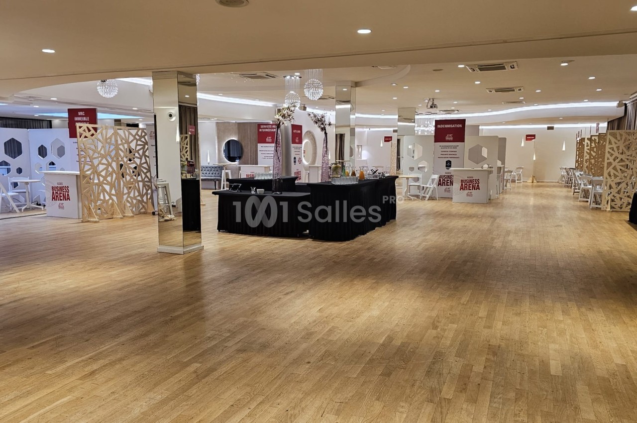 Salle spacieuse avec parquet clair, stands d'exposition, tables et chaises, éclairée par des plafonniers modernes.