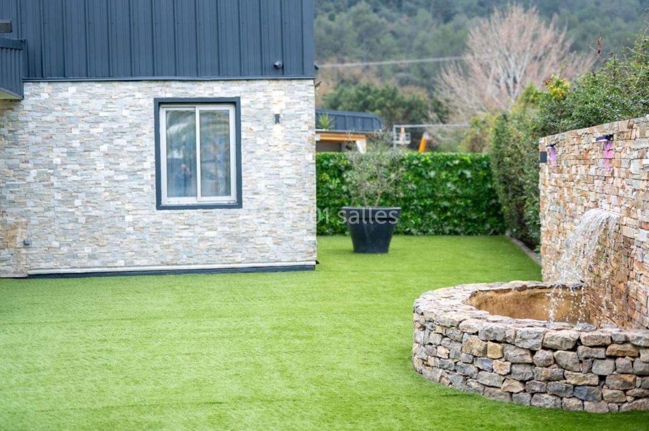 Jardin avec pelouse synthétique, mur en pierre, fontaine circulaire et haie de verdure en arrière-plan.