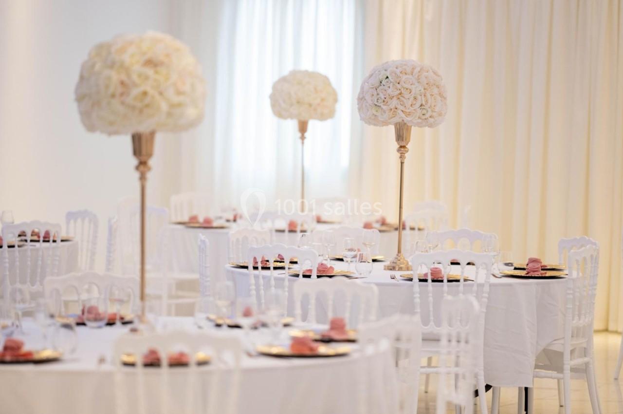 Salle de réception élégante avec tables rondes dressées, chaises blanches et centres de table ornés de fleurs blanches.