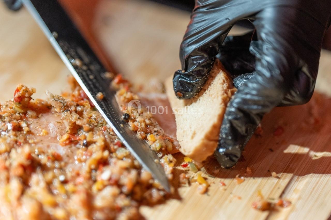 Une main gantée tient un morceau de pain près d'un couteau et d'une préparation hachée sur une planche en bois.