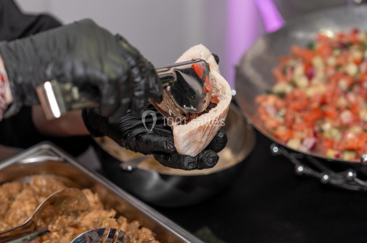 Une main gantée remplit un pain pita avec des légumes et de la viande, devant des plats garnis en arrière-plan.