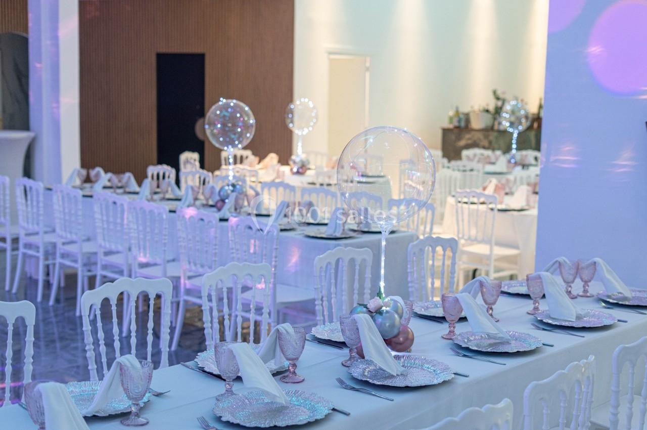 Salle de réception décorée avec des tables dressées, nappes blanches, assiettes élégantes et ballons lumineux.