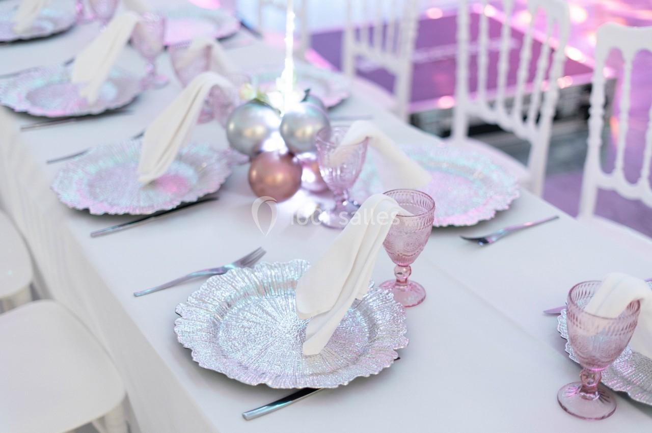 Table dressée avec des assiettes argentées, serviettes blanches, verres roses et décoration centrale de boules brillantes.