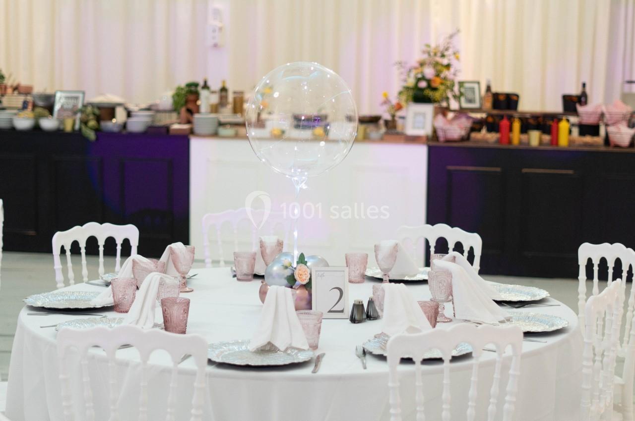 Table ronde élégamment dressée avec nappes blanches, vaisselle raffinée et un ballon transparent en centre de table.