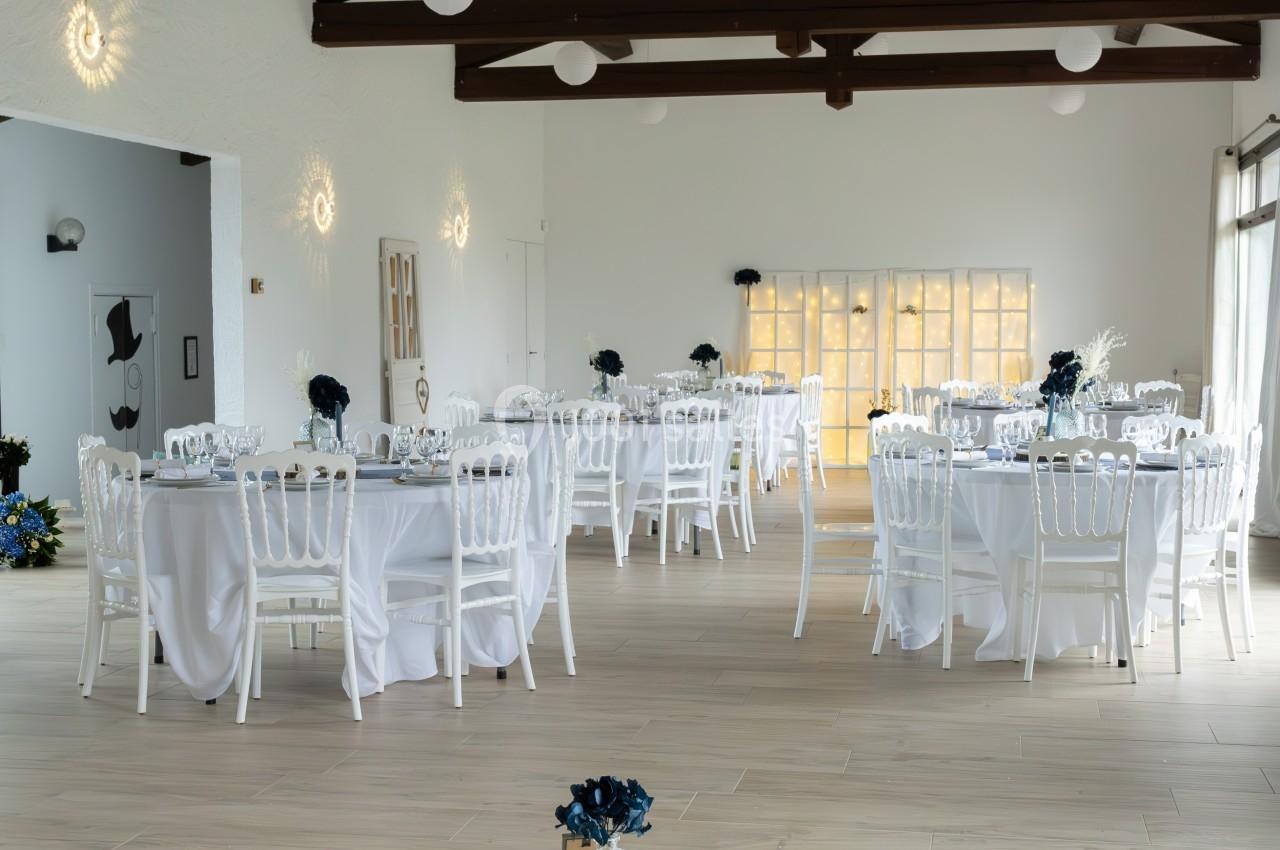 Salle de réception lumineuse avec tables dressées, chaises blanches et espace central décoré de coussins et vaisselle.