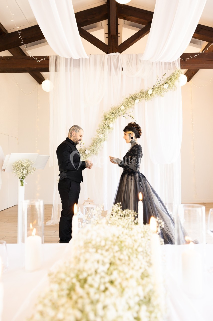 Un couple échange ses vœux dans une salle décorée de voilages blancs et de guirlandes florales.