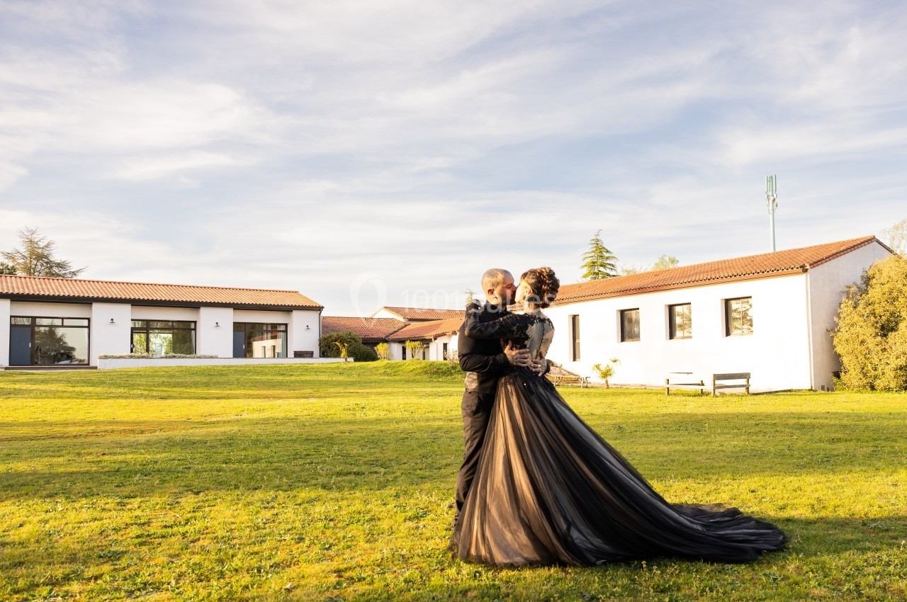 Un couple en tenue élégante s'enlace sur une pelouse devant des bâtiments modernes sous un ciel dégagé.