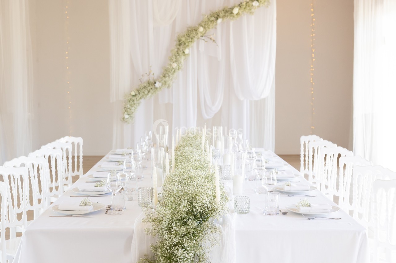 Salle décorée pour un événement, avec une longue table blanche ornée de gypsophile et un fond de voilages et guirlandes…