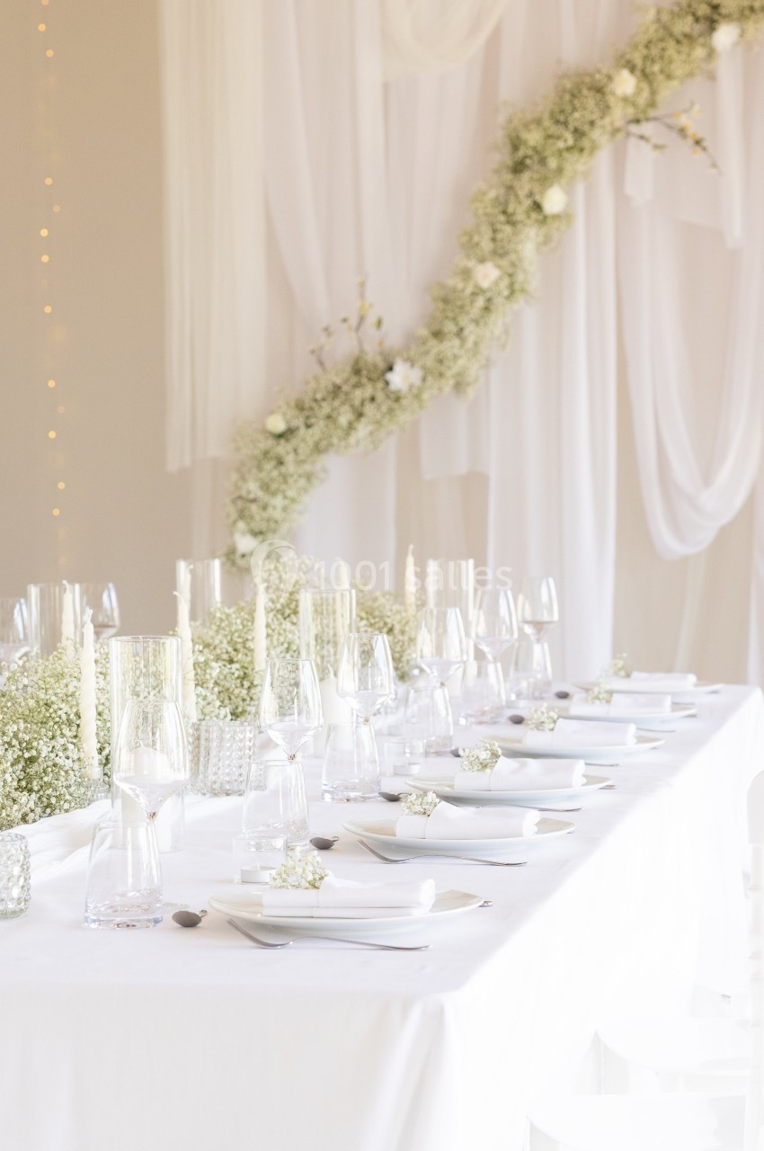 Table élégamment dressée avec nappes blanches, vaisselle raffinée et décoration florale blanche.
