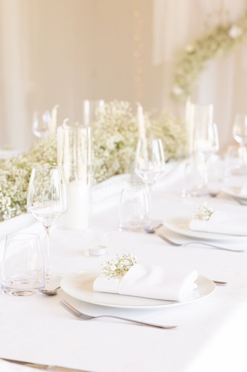 Table élégamment dressée avec nappes blanches, assiettes, verres, serviettes ornées de fleurs et centre de table floral.