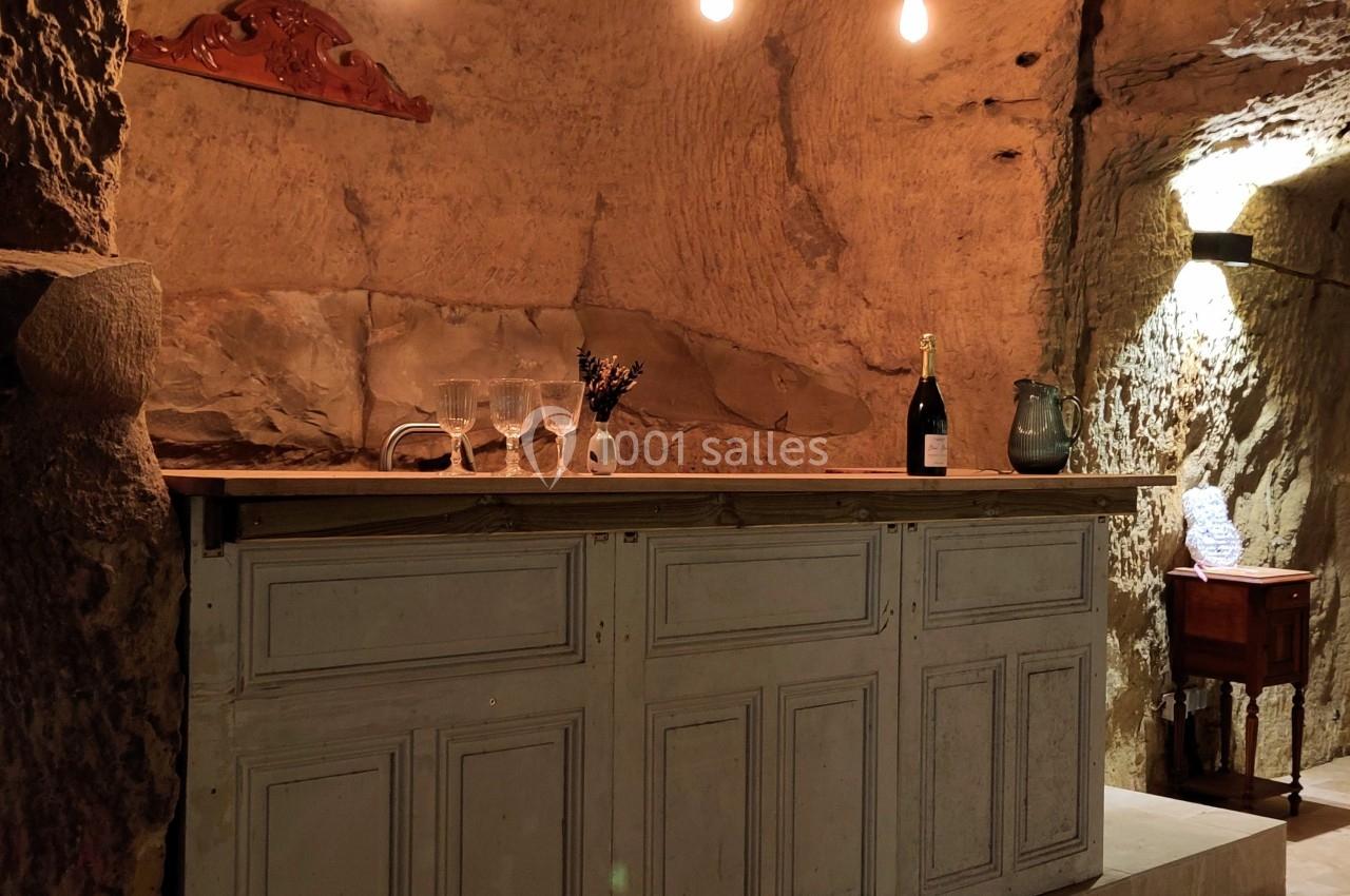 Comptoir en bois clair dans une cave en pierre éclairée par des ampoules suspendues, avec des verres et une bouteille.