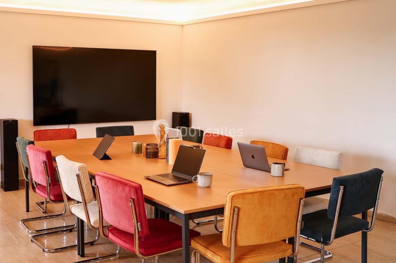 Salle de réunion avec une grande table en bois, des chaises colorées, des ordinateurs portables et un écran mural.