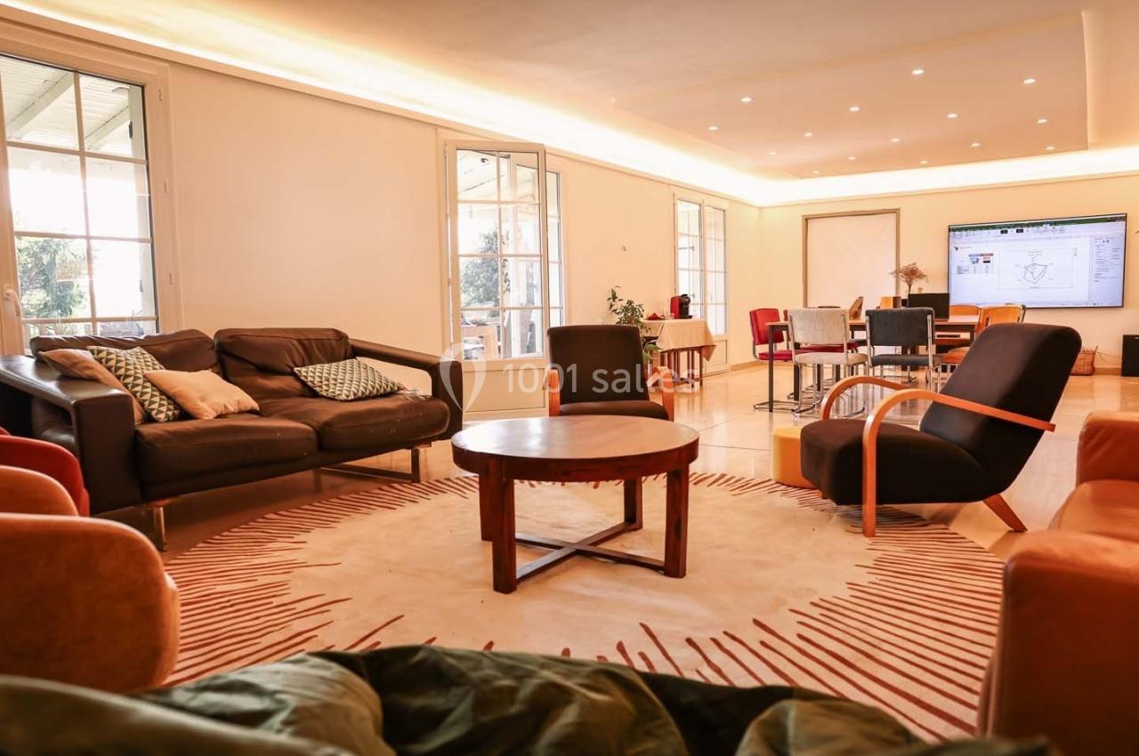 Salon lumineux avec canapés, fauteuils, table basse, tapis beige et écran affichant un plan technique en arrière-plan.