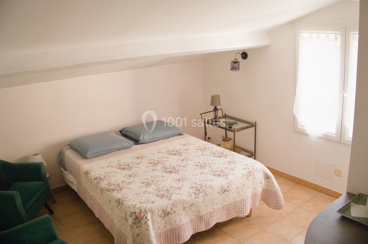 Chambre lumineuse sous combles avec lit double, fauteuil vert, table de chevet et fenêtre voilée.
