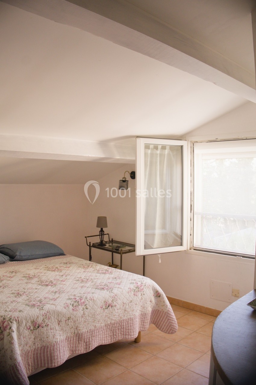 Chambre mansardée lumineuse avec lit double, fenêtre ouverte et table de chevet équipée d'une lampe.