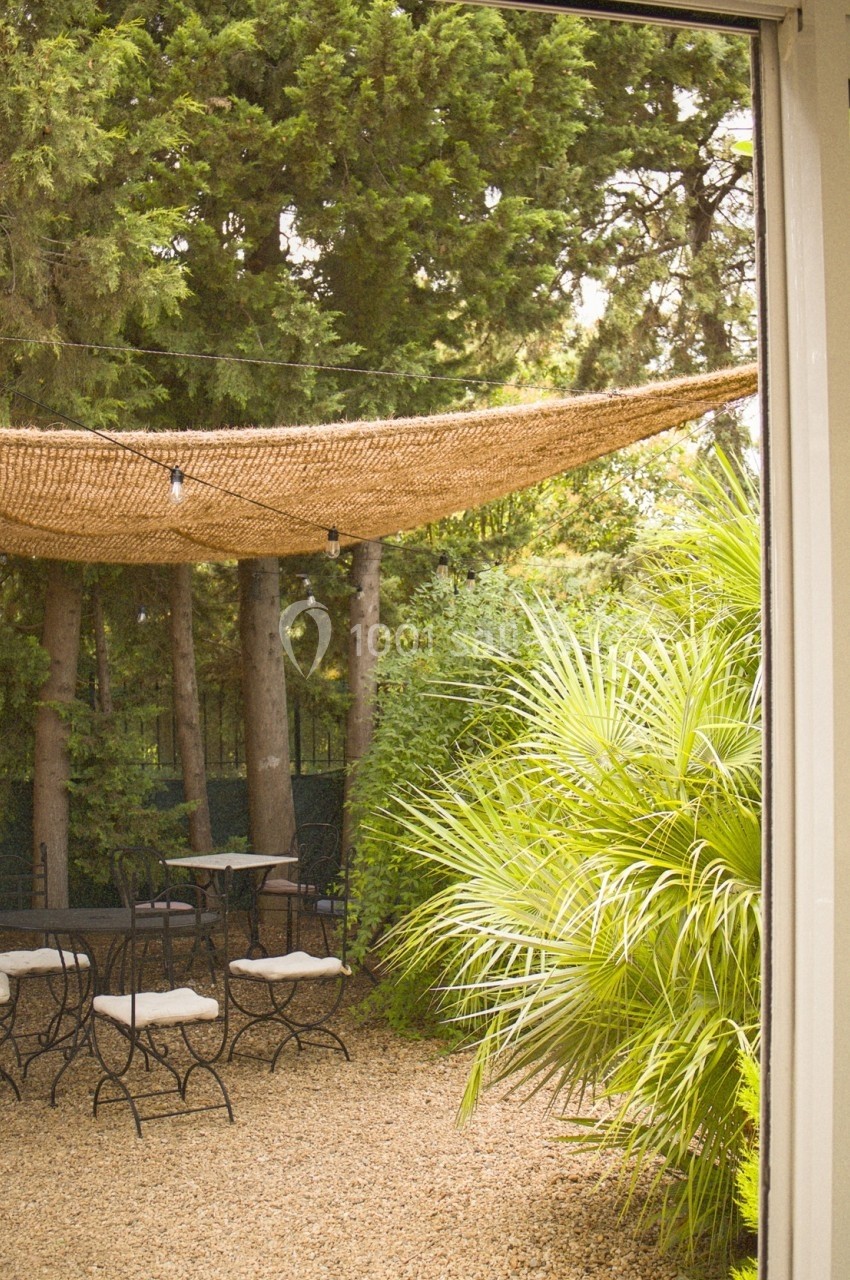 Vue d'une terrasse extérieure avec des tables et chaises, ombragée par une toile, entourée de végétation dense.